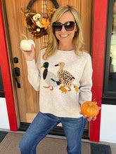 A woman wearing a cream-colored knit sweater with a bird and chick graphic, blue jeans, and sunglasses, posing in front of a wooden front door decorated with a fall wreath and holding a small pumpkin.