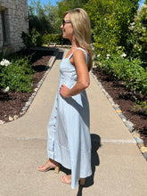Woman in a blue striped dress standing on a sidewalk with greenery in the background