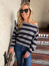 A woman wearing a gray and navy blue horizontally striped off-the-shoulder sweater, blue jeans, and black sunglasses, standing in a doorway with tiled steps in the background.