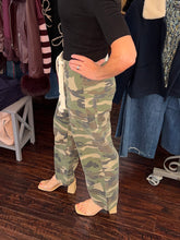 A person wearing loose camouflage cargo pants with a black top and beige block-heel sandals, standing in a clothing store on a dark wood floor with racks of clothes in the background.