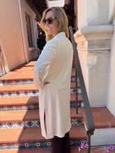 A woman wearing a long cream-colored coat and sunglasses stands on tiled outdoor stairs, looking back over her shoulder.