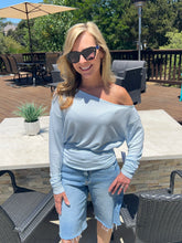 Woman wearing a light blue off-shoulder top and jeans standing outdoors on a patio.