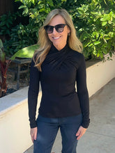 A woman wearing a black long-sleeve mock neck top and dark sunglasses, standing outdoors in front of green foliage and a beige wall.