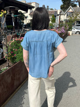 Woman wearing a chambray henley top and sunglasses standing outdoors with plants and buildings in the background.