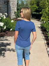 Woman wearing a blue t-shirt and jeans walking on a sidewalk with greenery and a stone building in the background.
