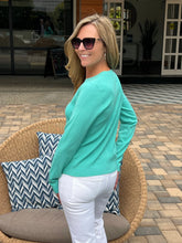 Woman in a teal sweater and white pants standing outdoors on a patio.