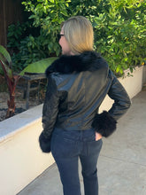 A woman wearing a black faux-leather jacket with a black fur collar and matching fur cuffs, shown from the back in an outdoor setting with green plants and a concrete walkway.