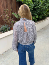 A woman viewed from behind wearing a gray and white abstract patterned long-sleeve blouse with a tie-neck and back cutout, paired with blue jeans, standing outdoors on a concrete patio with plants and a wooden fence in the background.