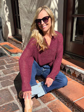 A woman wearing a textured burgundy knit long-sleeve V-neck sweater and blue jeans sits on terracotta steps outdoors in front of a building wall.