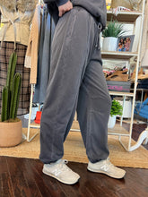 A person wearing loose, wide-leg gray sweatpants with drawstrings and light beige sneakers, standing in a retail or boutique interior with clothing racks, shelves, and plants visible.