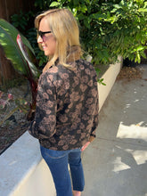 A woman wearing a brown and beige floral print barn jacket with blue jeans, and sunglasses, standing outdoors on a concrete walkway next to a garden.