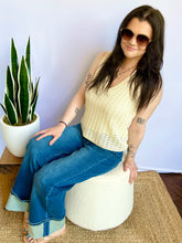 Woman wearing a cream sleeveless top and blue jeans standing next to a potted plant against a white wall.