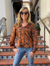 A woman wearing a rust-orange long-sleeve blouse with a dark abstract floral pattern and blue jeans, standing on tiled outdoor stairs.