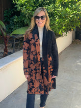 A woman wearing a long brown and black floral patterned coat over a cream turtleneck sweater and black pants, standing outdoors on a concrete walkway with green plants in the background.