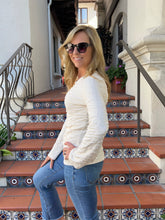 A woman wearing a cream ribbed long-sleeve top and blue jeans stands on tiled outdoor stairs in front of a building.