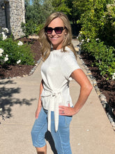 Woman wearing sunglasses and a white top with blue jeans standing on a sidewalk with greenery in the background.