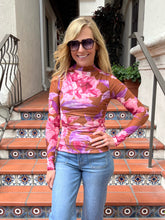 A woman wearing a long-sleeve mock neck top with a pink and purple floral pattern over a brown base, paired with blue jeans, standing on tiled outdoor stairs.