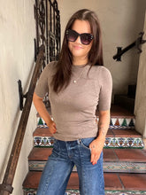 A woman wearing a fitted taupe short-sleeve knit top and blue jeans stands on a tiled staircase indoors. She is also wearing dark sunglasses and a delicate necklace.