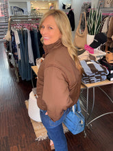 A woman with blonde hair wearing a brown puffer jacket and blue jeans, standing inside a clothing boutique and looking back over her shoulder. A blue handbag is hanging next to her.