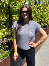 A woman wearing a textured light gray short-sleeve top with a chest pocket and black pants, standing outdoors in front of green plants and a wooden planter.