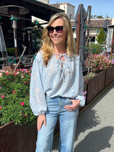 Woman wearing a light blue floral blouse and jeans standing in an outdoor setting with plants and buildings in the background.