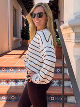 A woman wearing a cream and brown horizontally striped knit sweater and dark sunglasses, posing on outdoor terracotta tile stairs.