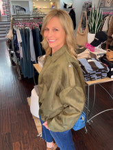 A woman with blonde hair smiling and looking back over her shoulder inside a clothing boutique. She is wearing an oversized olive green jacket and blue jeans, with a blue handbag hanging at her side.