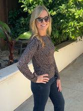 A woman wearing a brown and black patterned long-sleeve knit top and dark jeans, standing outdoors on a concrete walkway with green plants and a low white wall behind her.
