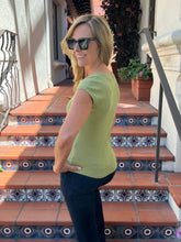 A woman wearing a short-sleeve olive green t-shirt and dark pants stands on outdoor tiled steps, looking back over her shoulder.