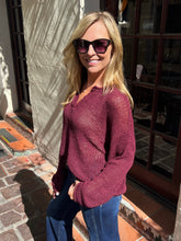 A woman wearing a maroon open-knit long-sleeve sweater and blue jeans, standing outdoors in front of a building with glass windows and tiled floors.