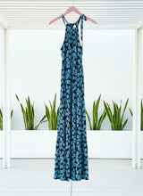 Blue medallion print maxi dress on a hanger against a white background with plants below.