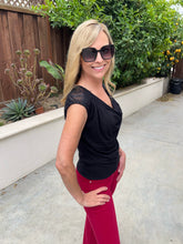A woman wearing a black short-sleeve lace-trim top and red pants, standing outdoors on a concrete walkway with a wooden fence and green plants in the background.