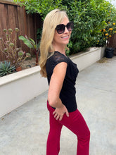 A woman posing outdoors in a black short-sleeve top with sheer lace detailing on the shoulders and bright red flare pants, wearing large black sunglasses.