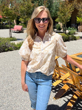 Woman wearing sunglasses and a patterned blouse standing outdoors with wooden chairs and greenery in the background.