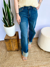 Person wearing blue jeans standing next to a plant and wooden box on a textured floor.