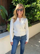 A woman wearing a cream cable-knit button-up cardigan with a small embroidered polo player logo on the chest, paired with blue jeans and sunglasses, standing outdoors in front of a beige wall and green foliage.