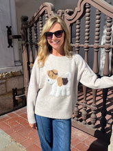 A woman wearing a cream-colored knit sweater with a dog motif and blue jeans, standing outdoors in front of a decorative brown metal railing.