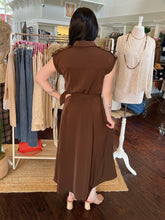 A woman stands with her back to the camera wearing a brown sleeveless midi dress with a collar and tie waist, inside a clothing boutique. Clothing racks and a mannequin are visible in the background.