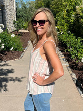 Woman wearing sunglasses and a striped tank top on a sidewalk with greenery in the background