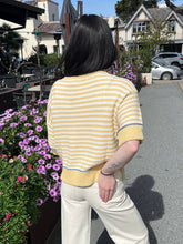 Person wearing a yellow striped sweater and white pants standing outdoors with flowers and buildings in the background.