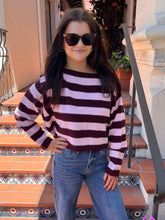 A woman wearing a long-sleeve knit sweater with horizontal dark brown and light pink stripes, paired with blue jeans and sunglasses, standing on outdoor tiled steps.