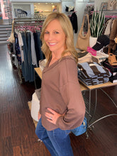 A smiling blonde woman wearing a brown ribbed long-sleeve top and blue jeans stands inside a clothing boutique, with racks of garments, folded sweaters, and store fixtures visible in the background.