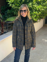 A woman wearing a brown and green plaid button-up coat over a black turtleneck and dark jeans, standing outdoors on a concrete walkway with green foliage in the background.