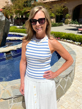 Woman in a striped top and white skirt standing by a pool with sunglasses on.