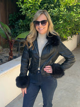 A woman wearing a black faux-leather moto jacket with dark faux-fur trim on the collar and cuffs, styled with dark jeans and sunglasses, standing outdoors on a concrete walkway with green foliage in the background.