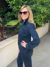 A woman wearing a dark blue denim jacket and matching dark jeans, standing outdoors on a concrete walkway with green foliage and a low beige wall behind her.