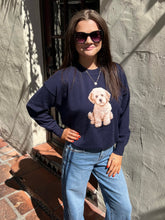 A woman wearing a navy blue crewneck sweatshirt with a graphic of a fluffy cream-colored dog, paired with light blue jeans, standing outdoors against a textured stucco wall.