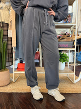 A person wearing dark gray drawstring sweatpants and light beige sneakers, standing in a retail store setting with clothing racks and shelves in the background.