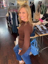 A woman posing in a clothing boutique, wearing a fitted brown mock neck long-sleeve top and blue jeans.