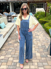 Woman in blue overalls standing on a patio with outdoor furniture and greenery.